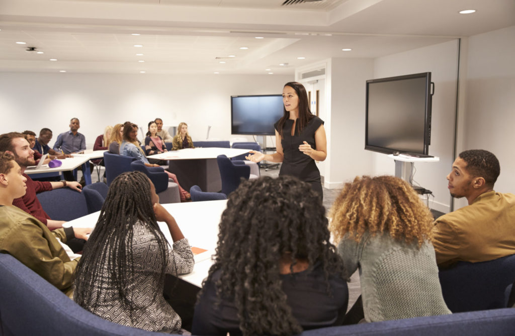 a woman lecturing to other people in a classroom with a monitor behind her