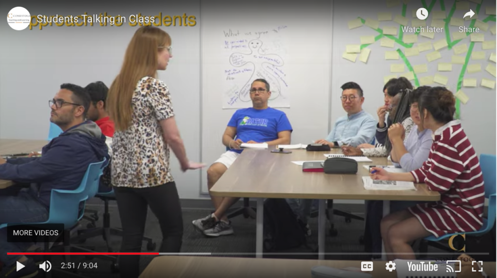 Screenshot of video, Students Talking in Class. Teacher approaching a table of students that are talking in class