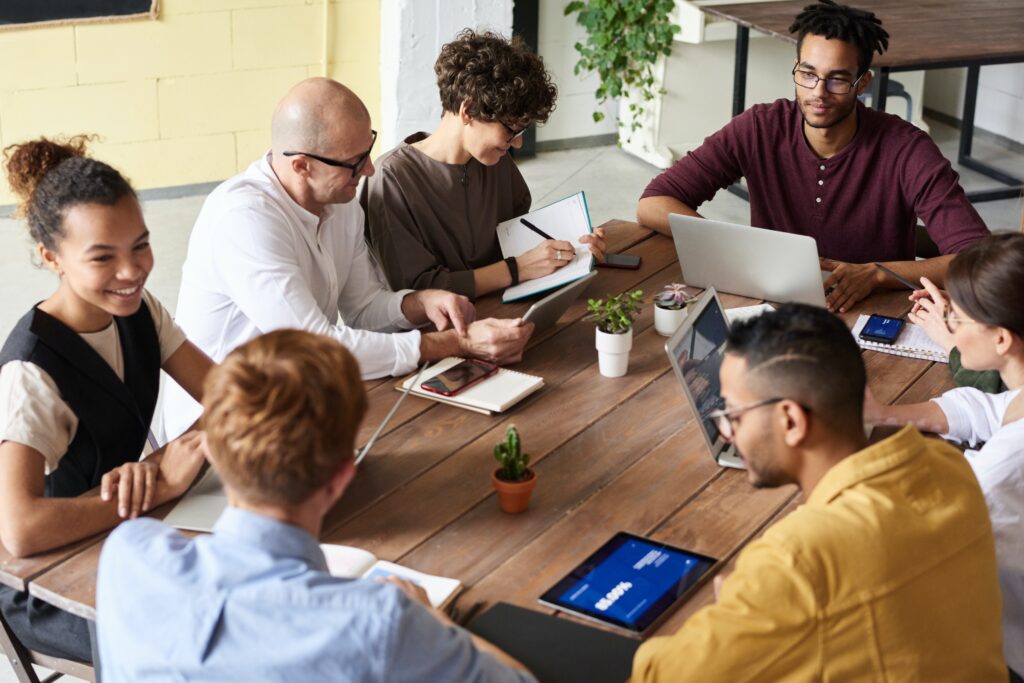 A group of adult learners holding notebooks and typing on computers and discussing with one another at a table