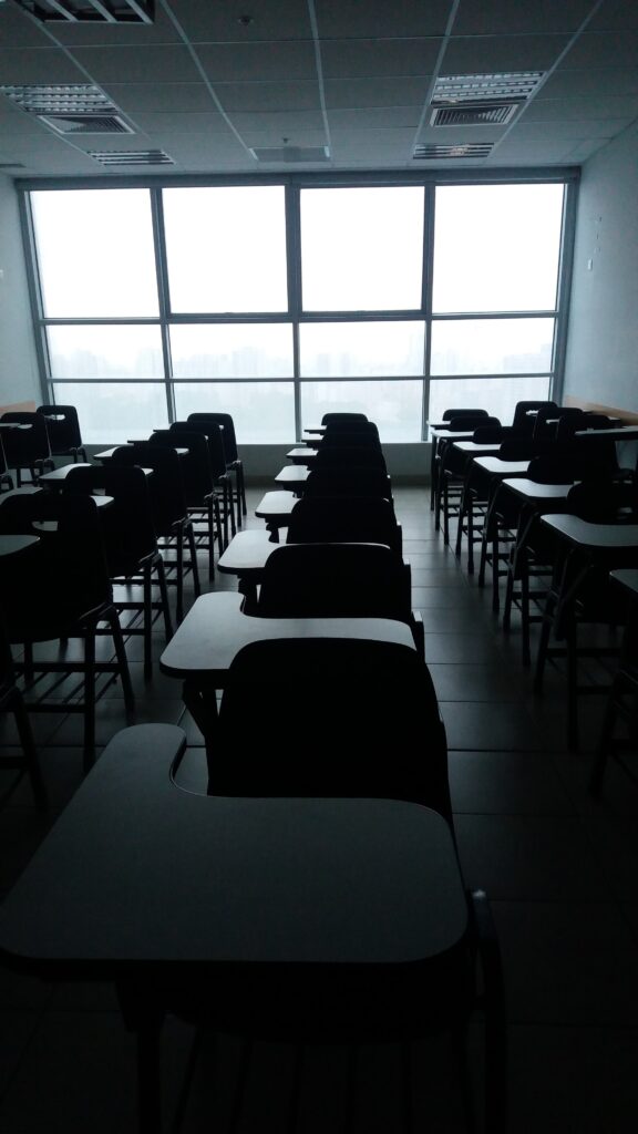 A classroom with the lights turned off, lit only by the large window alongside rows of desks.