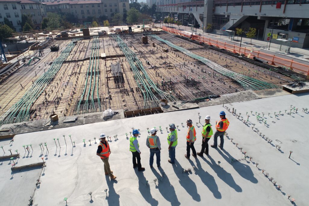 A group of construction workers stand on a rooftop examining blueprints and deciding what next steps they should take to finish a building.