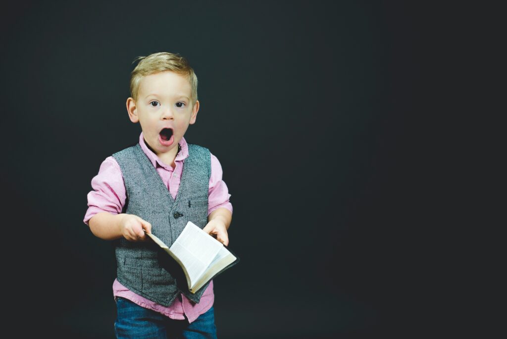 a little boy in a blue vest and pink shirt holding a book and looking suprised