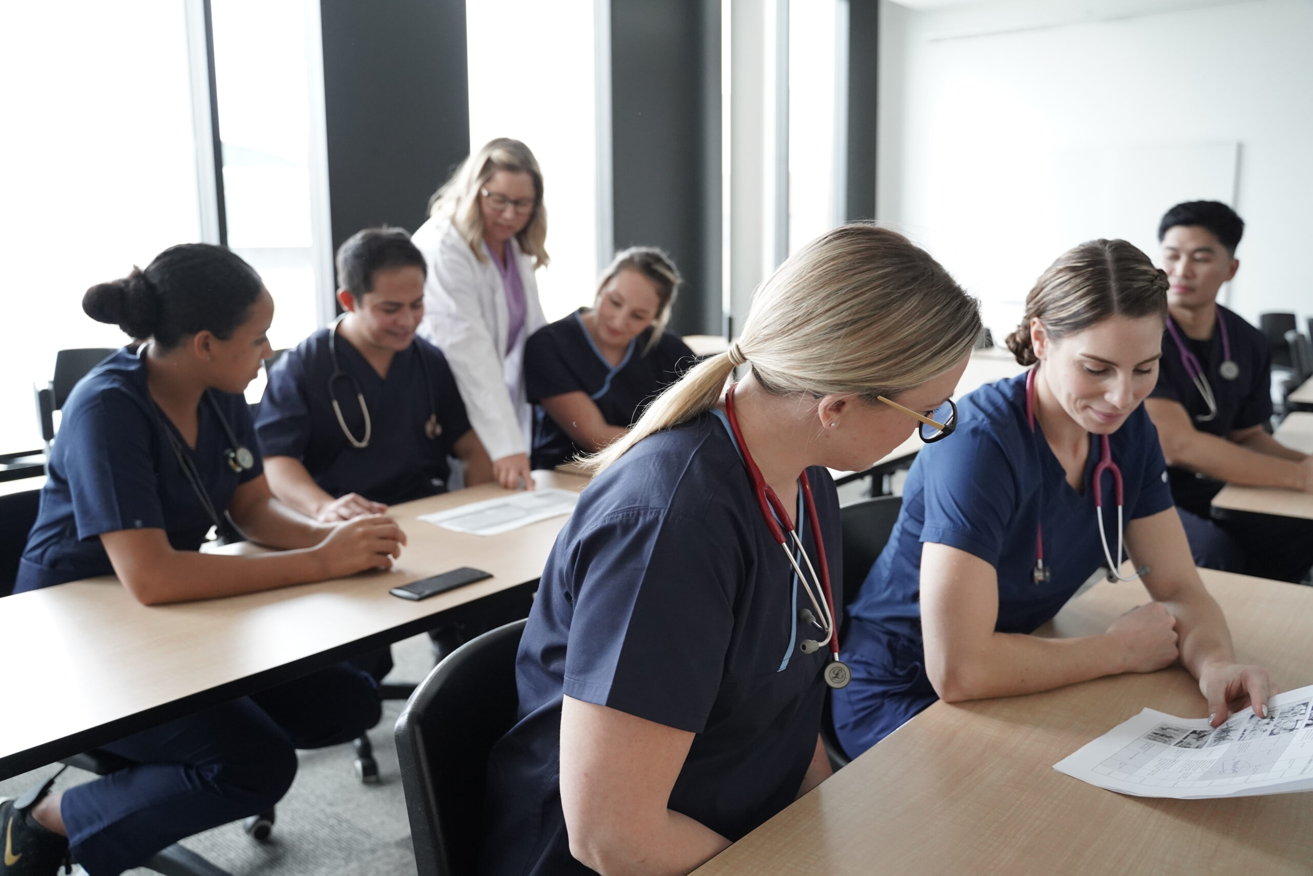 Nursing students wearing scrubs and stethoscopes, sit in small groups at wooden tables as they review worksheets together. An educator assist the group in the back with their work.