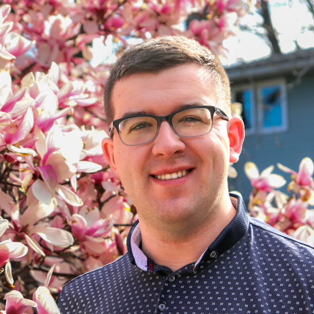 A smiling man with brown hair standing just in front of magnolia tree branches in full bloom, with a building tucked away in the background