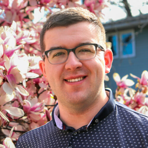 A smiling man with brown hair standing just in front of magnolia tree branches in full bloom, with a building tucked away in the background