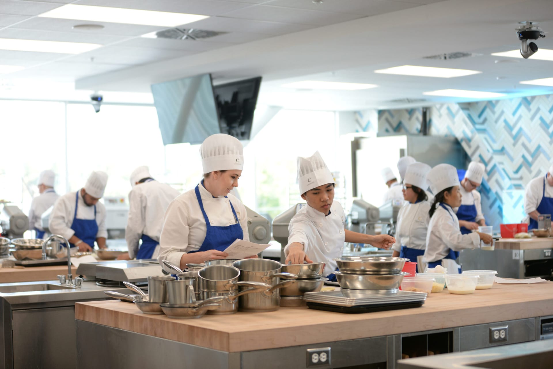 A class of Conestoga culinary students in their white cooking attire and aprons, working with pots and pans at various food preparation stations.