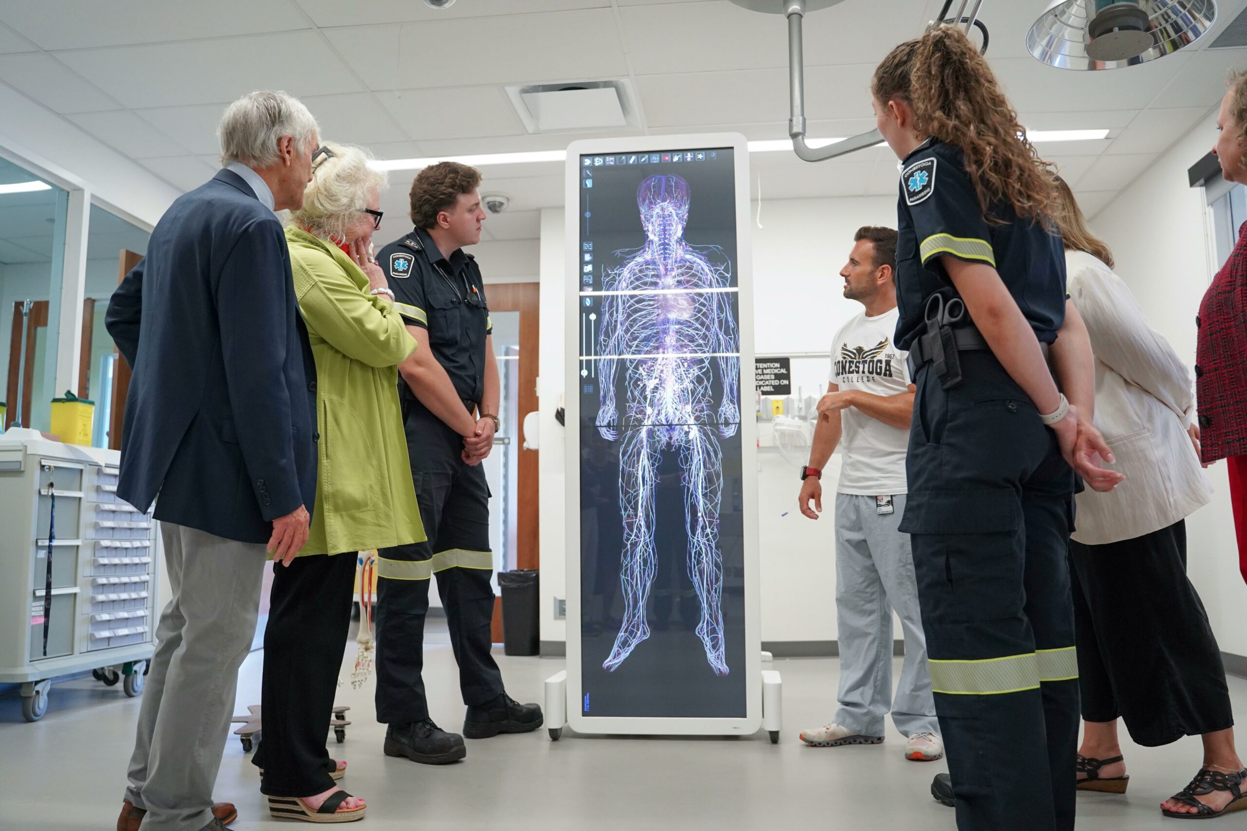 Conestoga staff and paramedics reviewing the output of a large body scan simulation machine called an Anatomage table. The table is upright appears to display an interactive virtual body's nervous and cardiovascular systems.