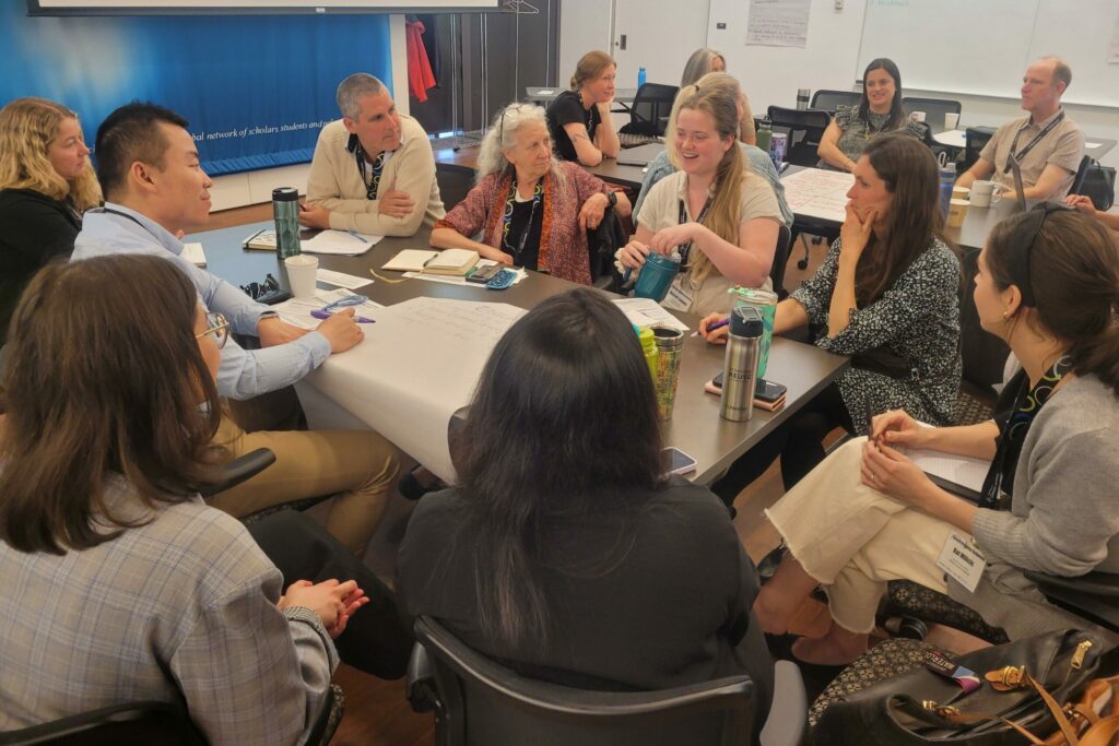People sit around long wooden tables, sharing ideas and writing them down on large sheets of paper at a break-out workshop at the Climate Pedagogy Symposium.