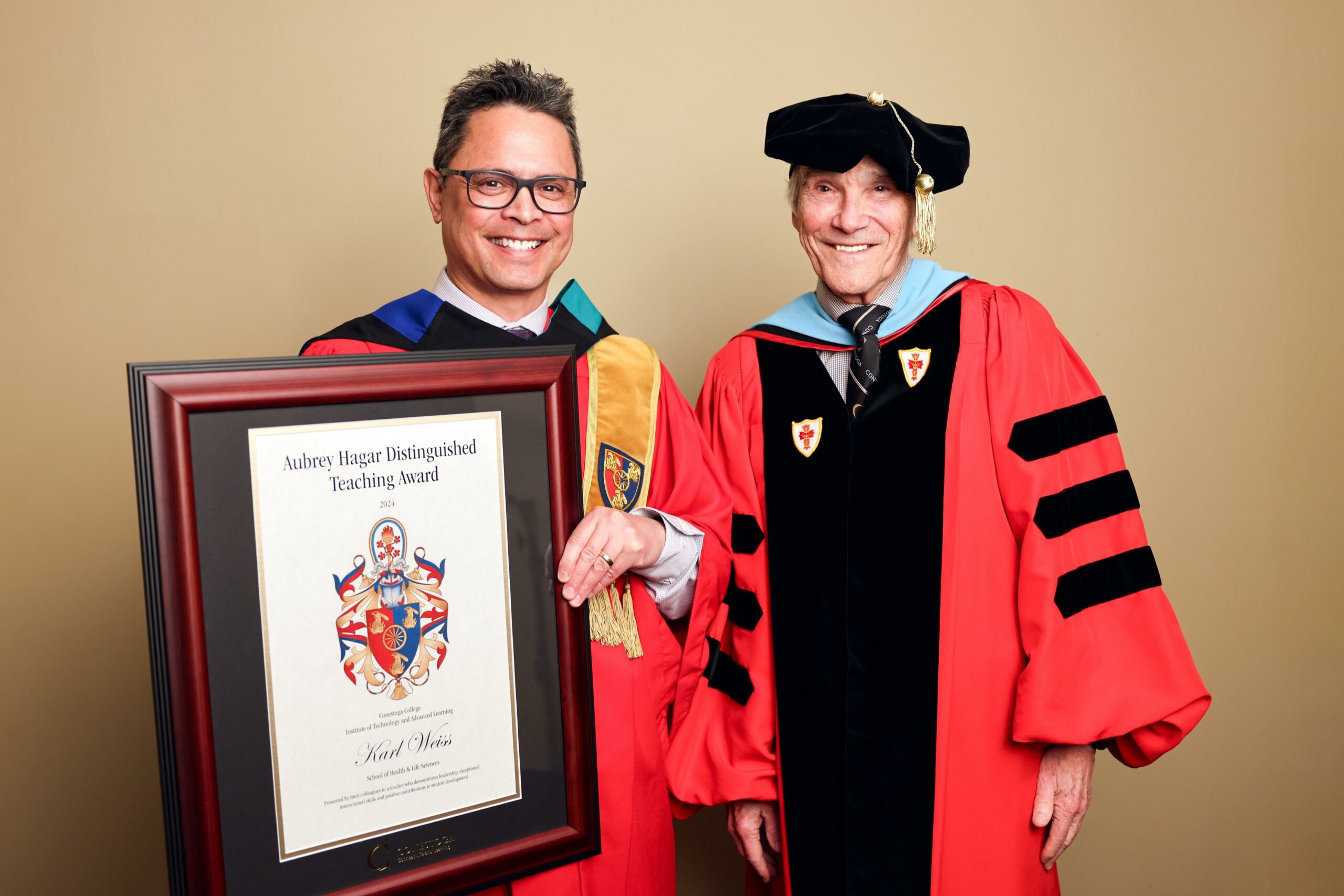 Phot of a man named Karl Weiss with square glasses posing with a large framed 2024 Aubrey Hagar Distinguished Teaching Award and standing in graduation robes beside a man in similar robes and a graduation cap, President John Tibbits, as part of the college's winter convocation celebration.